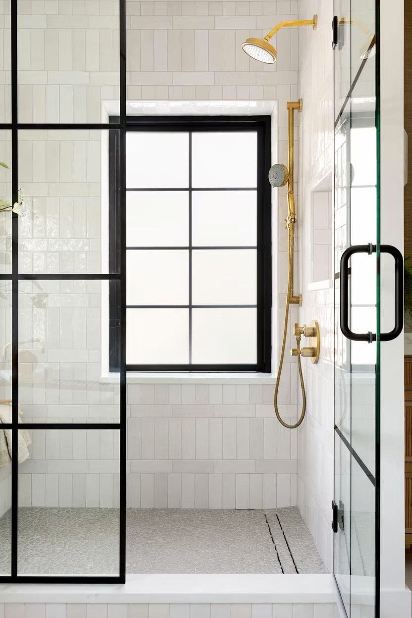 White vertical tile shower with a black-framed window, brass fixtures and a pebble floor
